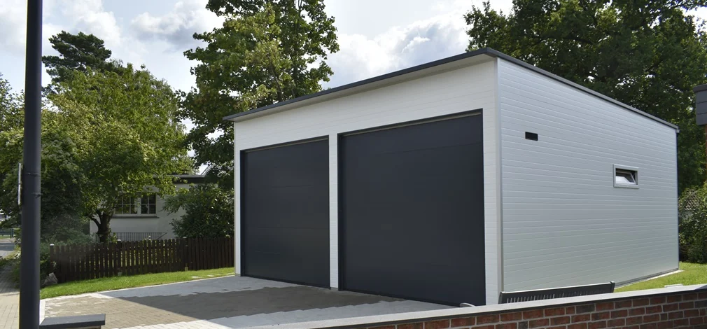 Doppelwohnmobilgarage aus Sandwichpaneelen mit zwei großen, anthrazitfarbenen Sektionaltoren und weißer Fassade. Großzügige Doppelgarage für Wohnmobile mit isolierter Sandwichpaneel-Konstruktion und modernem Flachdach. Isolierte Doppelgarage mit zwei Sektionaltoren in Anthrazit und seitlichem Fenster für Tageslicht. Hochwertige Doppelwohnmobilgarage aus Sandwichpaneelen – langlebig, energieeffizient und pflegeleicht. Moderne Doppelgarage für Wohnmobile mit weißer Sandwichpaneel-Fassade und anthrazitfarbenen Toren