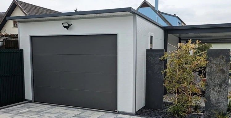 Moderne Fertiggarage aus Sandwichpaneelen mit anthrazitfarbenem Sektionaltor und Flachdach – hochwertige und isolierte Garagenlösung. Isolierte Garage mit Sandwichpaneel-Fassade, glattem Sektionaltor in Anthrazit und angrenzendem Carport. Hochwertige Fertiggarage mit Wärmedämmung, Sandwichpaneel-Wänden und modernem Flachdachdesign. Sandwichpaneel-Garage mit pflegeleichter Fassade, isoliertem Sektionaltor und eleganter Hofeinfahrt. Freistehende Garage mit Sandwichpaneel-Konstruktion und Sektionaltor in zeitlosem Anthrazitgrau