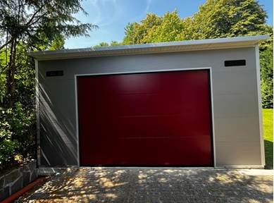 Freistehende Einzelgarage in Sonderanfertigung aus wärmedämmenden Sandwichpaneelen mit grauer Außenfassade und rotem Sektionaltor – moderne und individuelle Lösung für Pkw