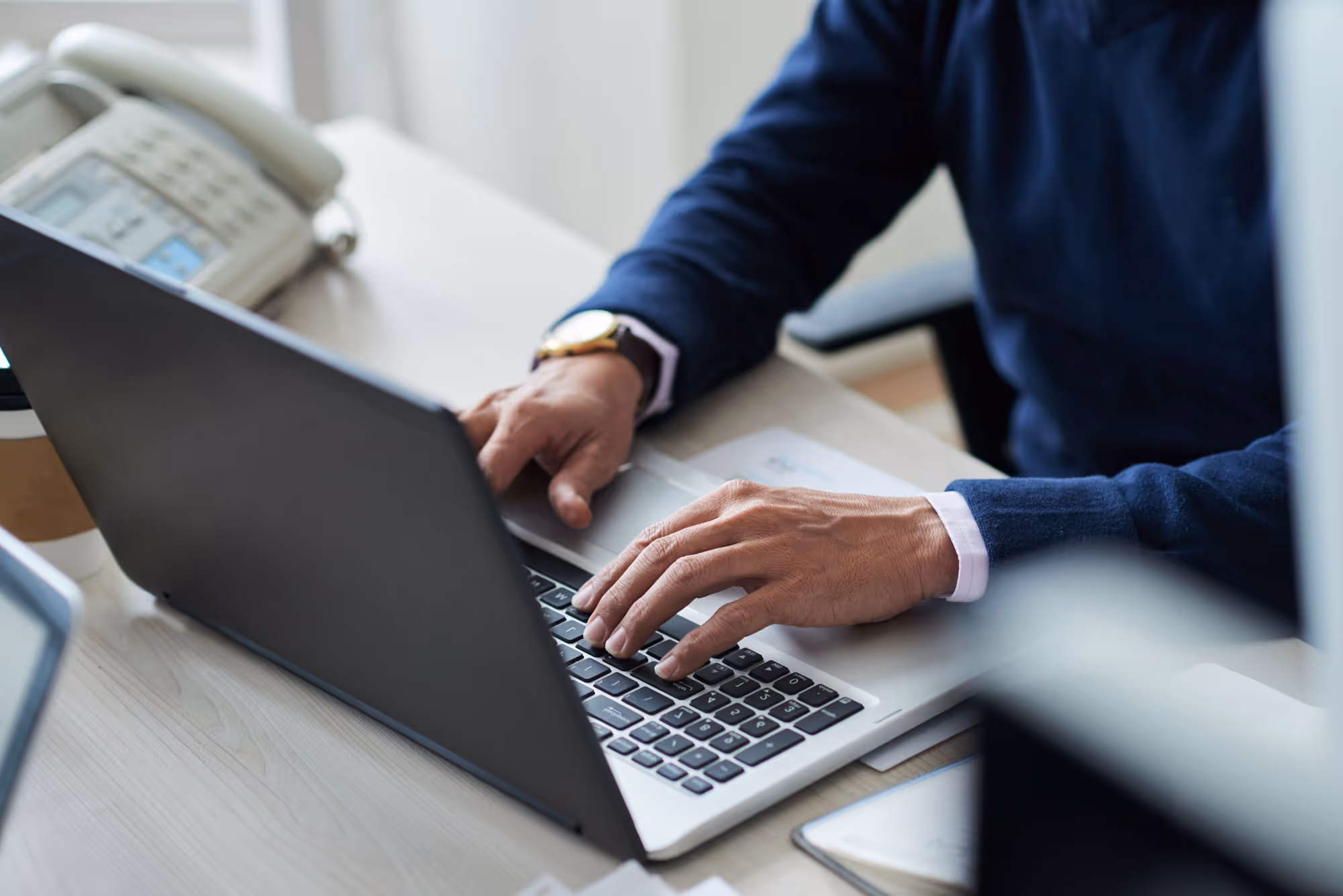 Person wearing a dark sweater typing on a laptop keyboard at a desk with a phone in the background.