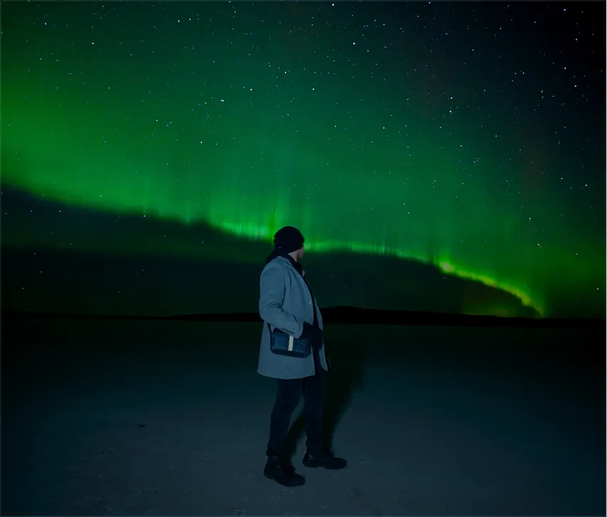 Man standing under vivid green northern lights on a snowy plain.