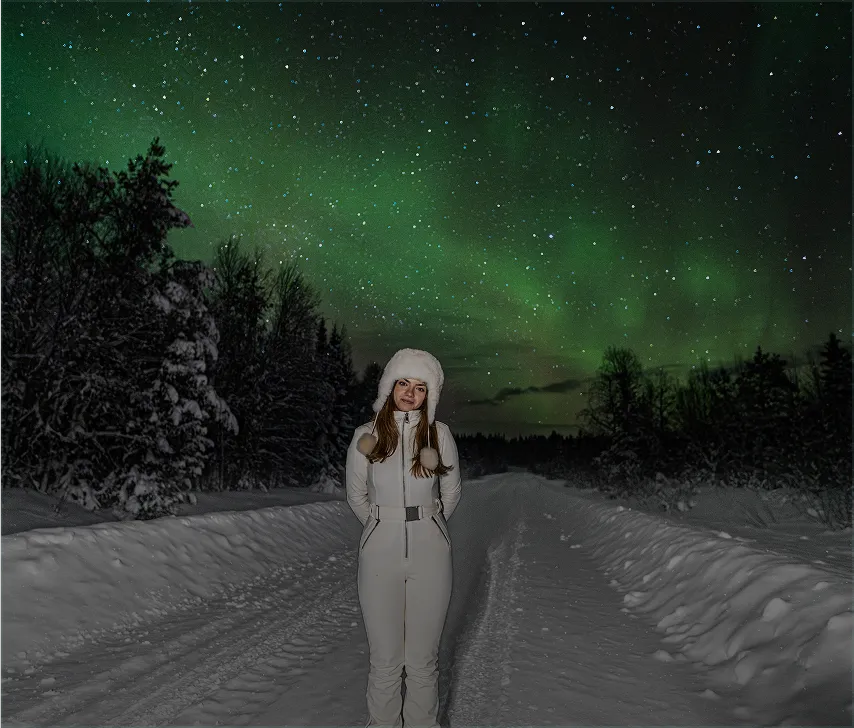 Woman in white winter outfit posing under northern lights on a snowy forest road.