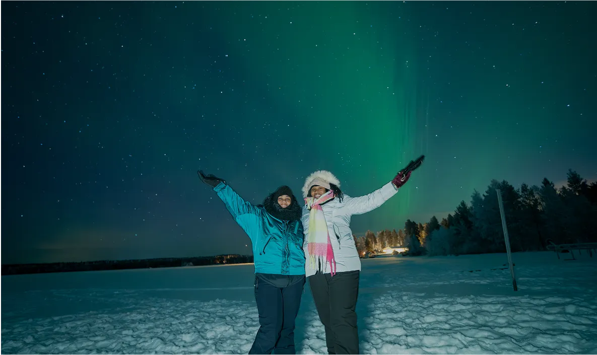 Two women posing with arms raised under green aurora.