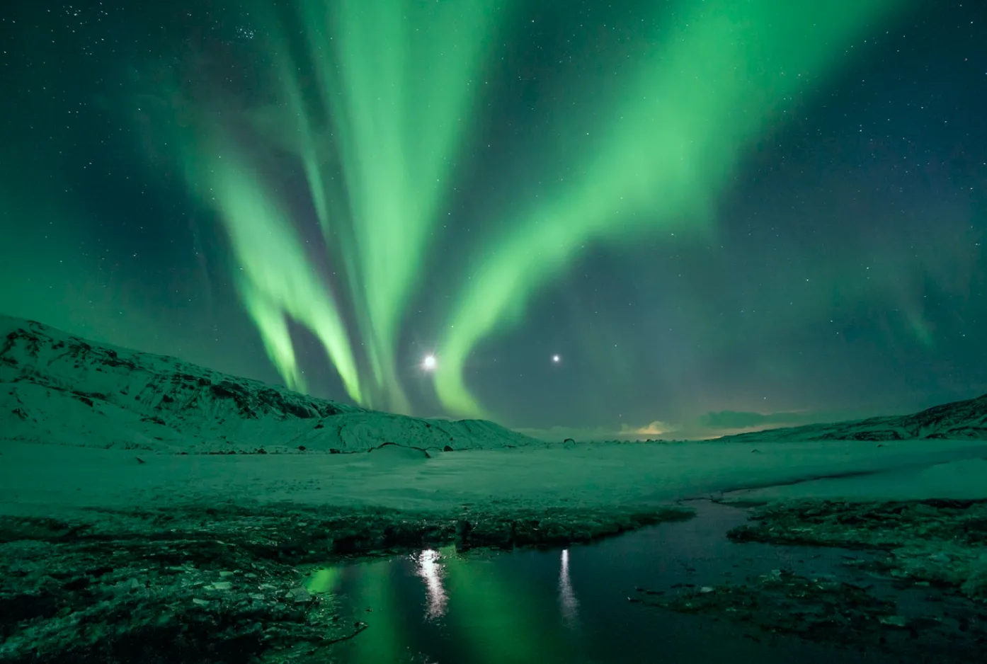 Bright green aurora dancing above a snowy mountain landscape with a reflective stream.