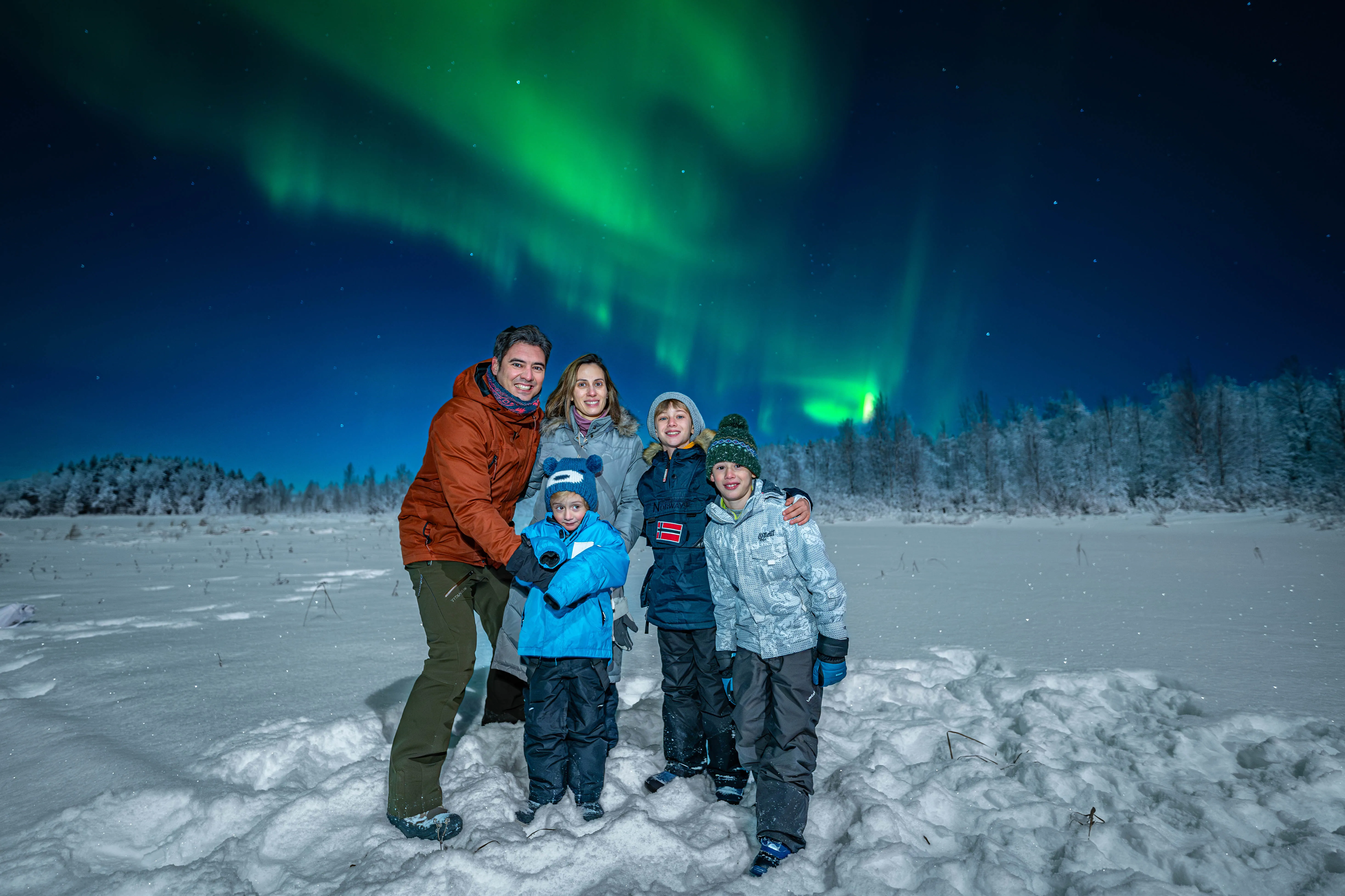 Smiling family standing on snowy field under vibrant green Northern Lights in Lapland.