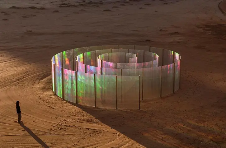 A solitary figure stands before a colorful, iridescent spiral installation in a vast, rocky desert landscape at sunset, conveying solitude and wonder.