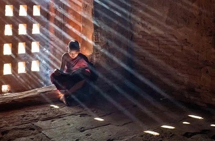 A young monk sits peacefully in a dimly lit room, bathed in dramatic sunlight streaming through a latticed window. The ambience feels tranquil and contemplative.