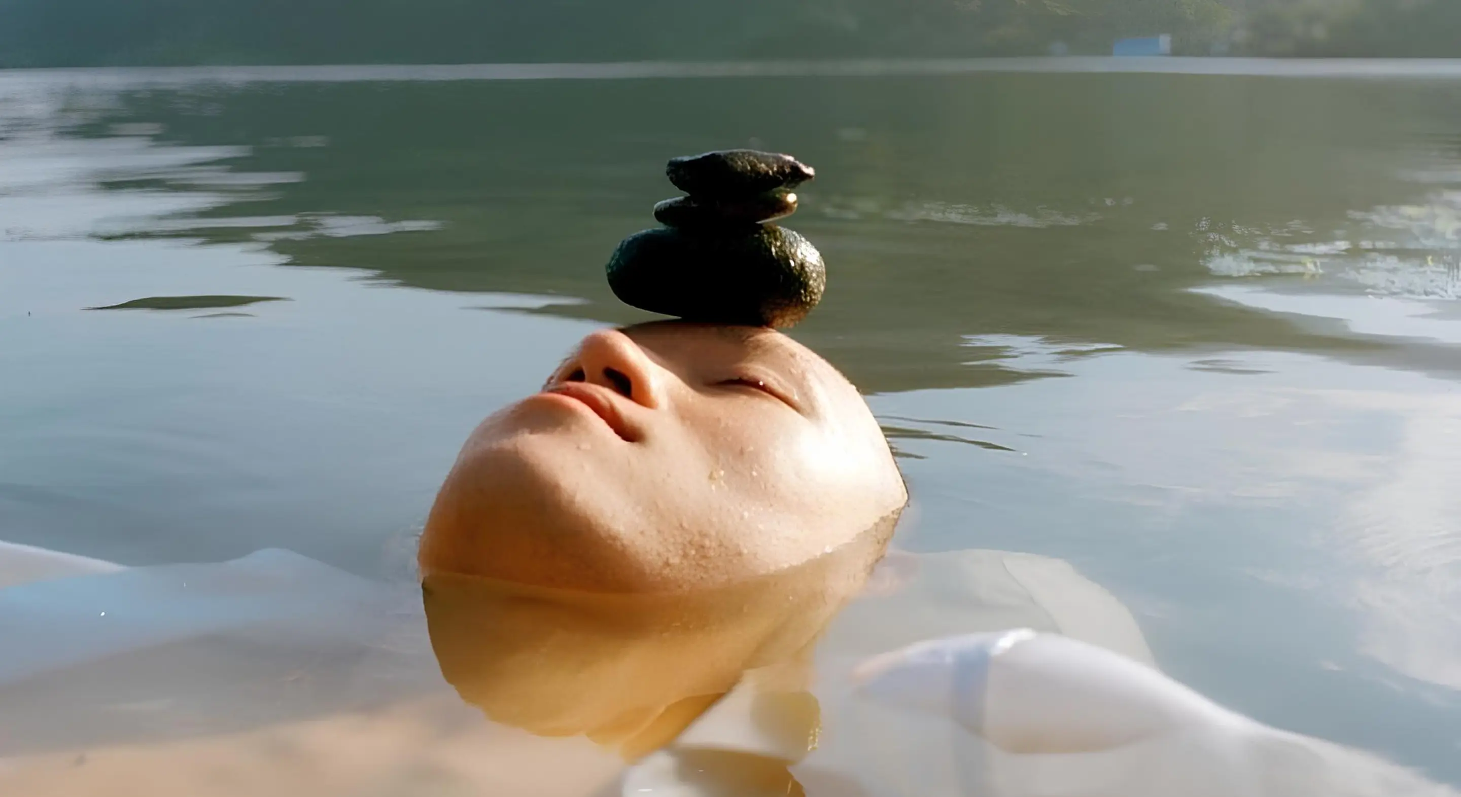 A serene face floats in calm water with eyes closed. Three small stones balance on the forehead.