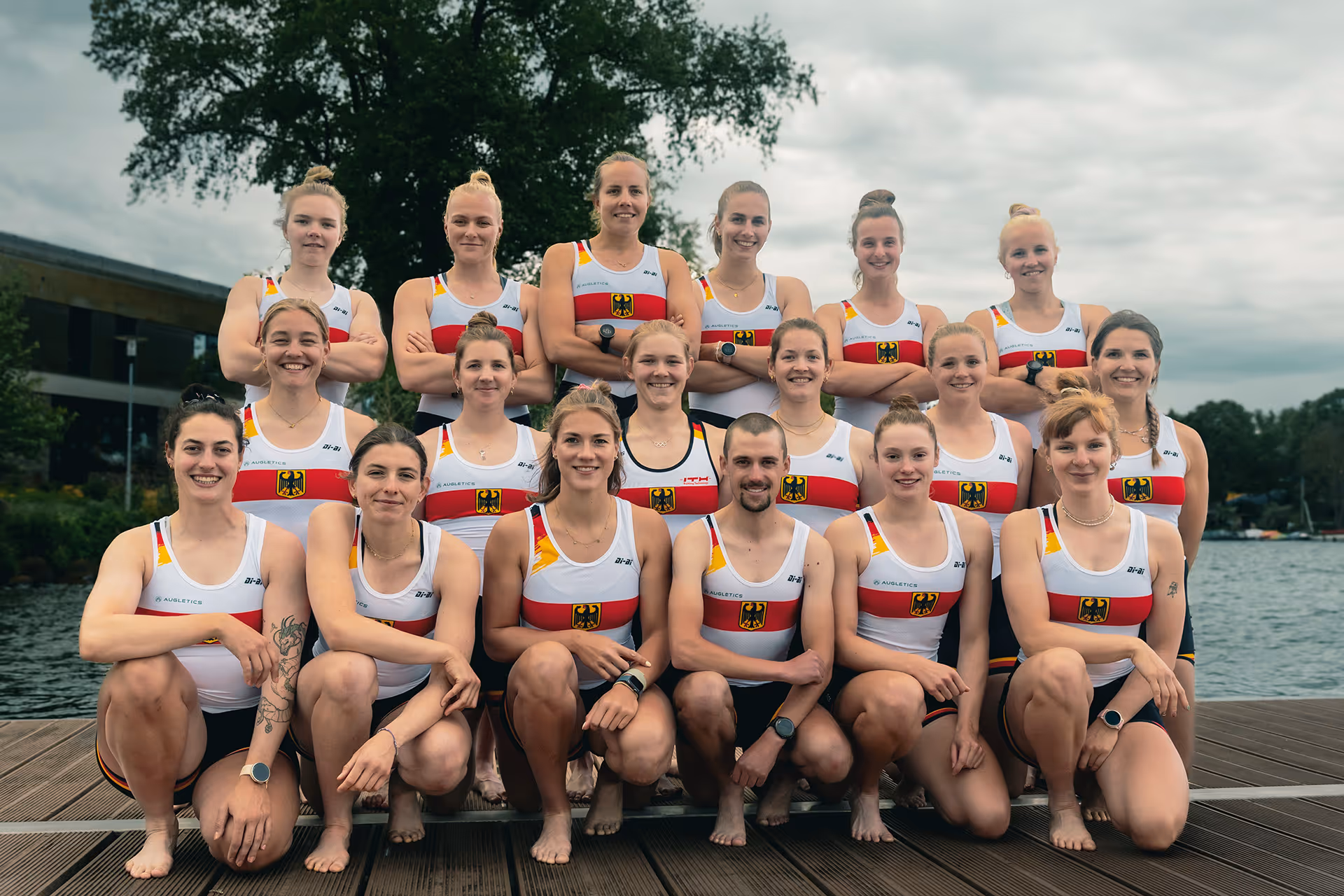 Gruppenfoto von 16 Athletinnen und einem Athleten im Einteiler des deutschen Nationalteams auf einem Steg am Wasser.