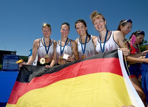 Vier Ruderinnen im Trainingsoutfit des deutschen Teams stehen mit einer Deutschlandfahne auf dem Siegersteg und tragen Goldmedaillen um den Hals