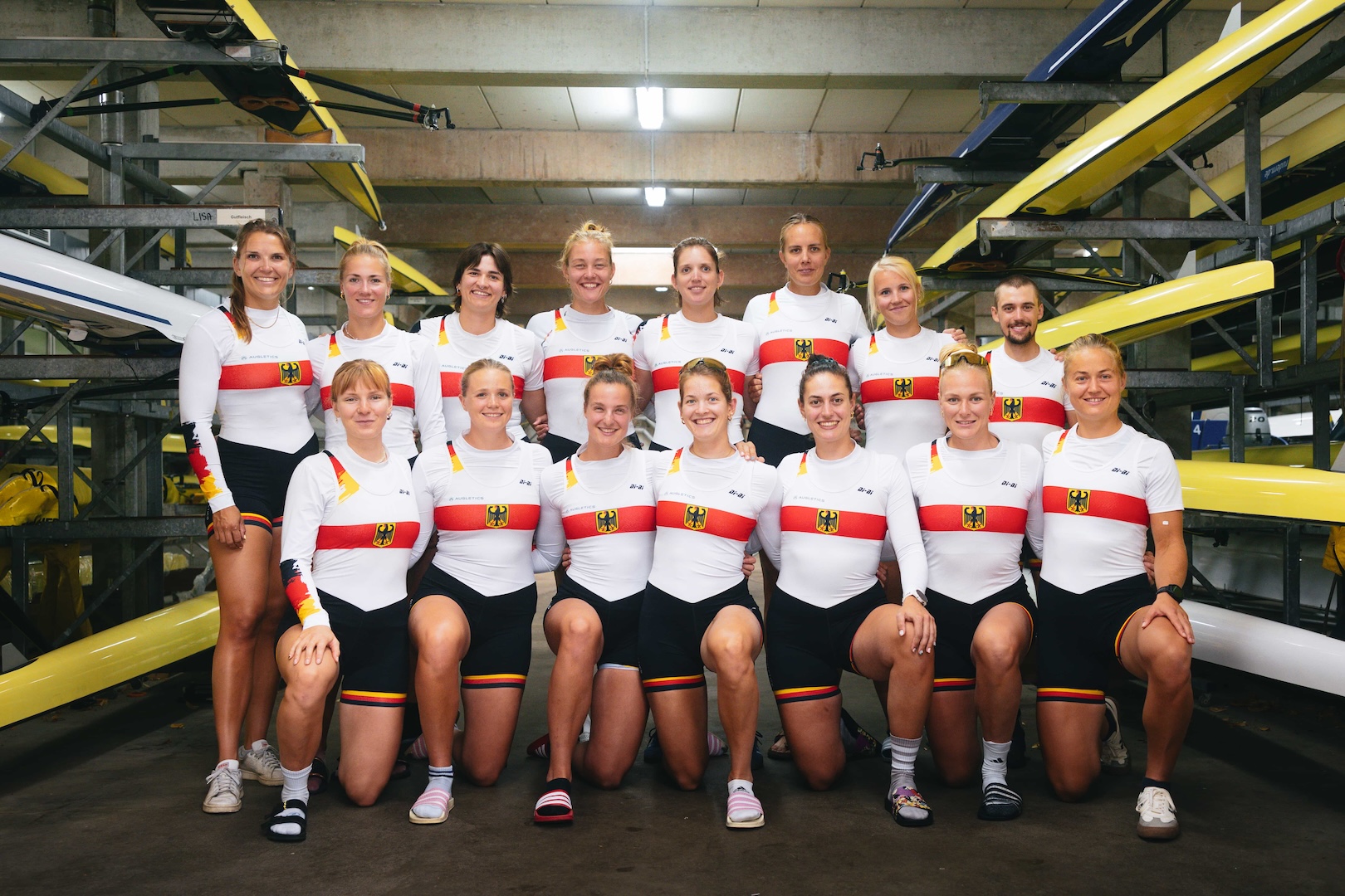 14 Ruderinnen und ein Steuermann im Trainingsoutfit des deutschen Teams stehen in der Bootshalle und posieren in die Kamera