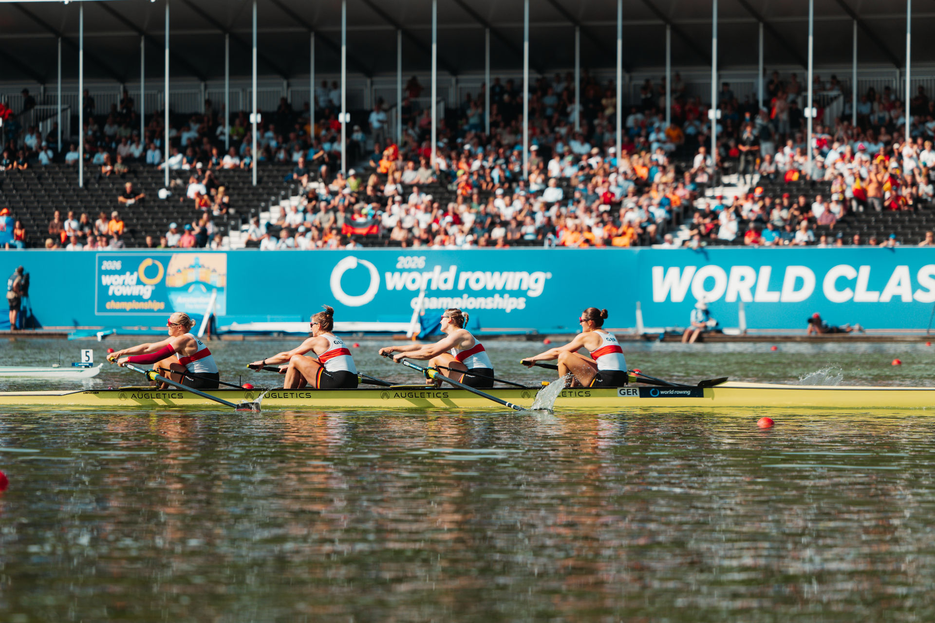 4 Ruderinnen rudern in einem Boot vor einer vollen Tribüne