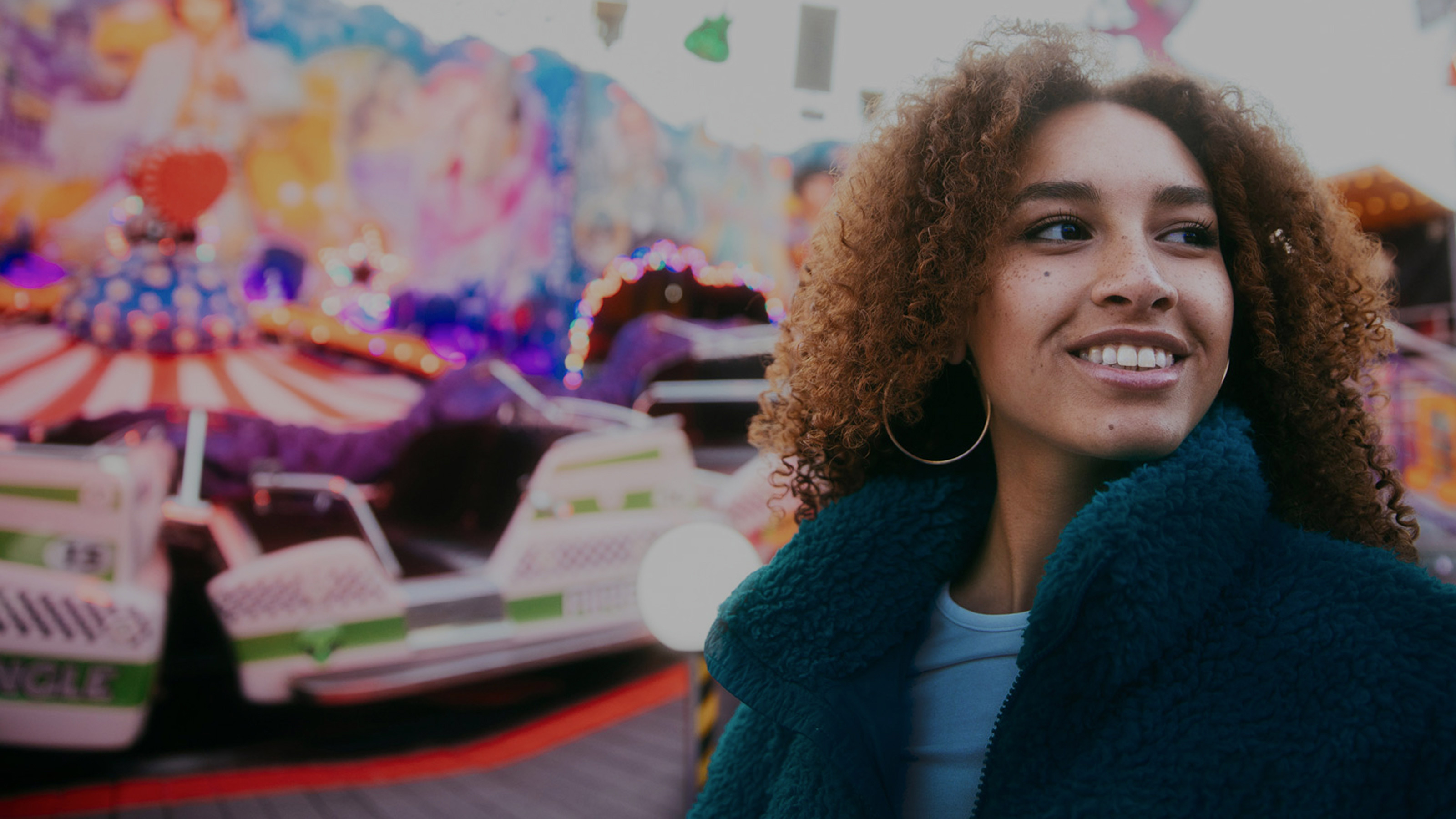A young woman with curly hair and hoop earrings smiles while standing in front of a colorful amusement ride. She wears a teal fleece jacket and a light blue top. The background is lively and blurred, filled with bright lights and vibrant painted panels, creating a fun, nostalgic fairground vibe.