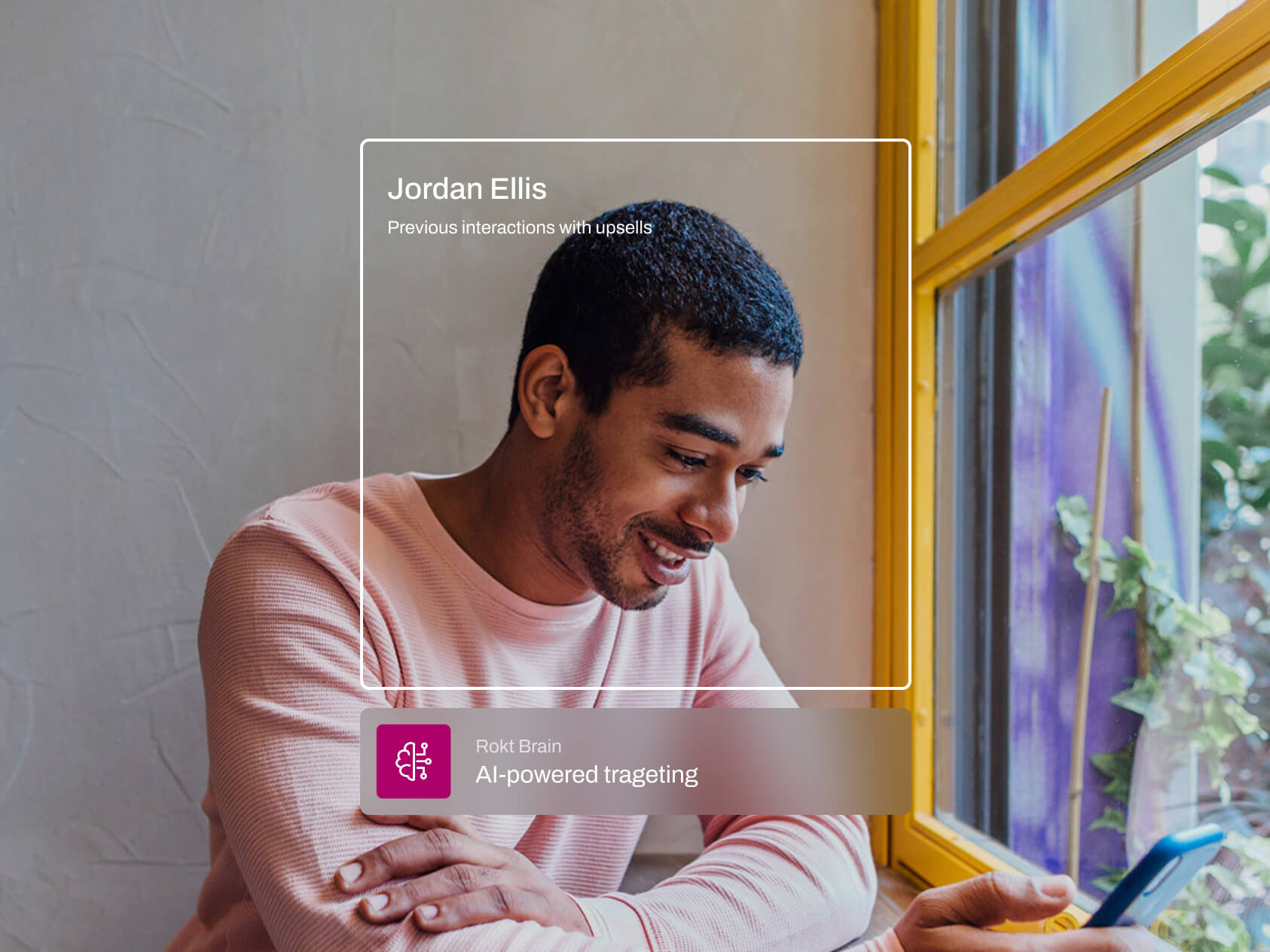 A man in a pink sweater sits at a café table by a bright yellow window, smiling as he looks at his phone. A floating frame identifies him as “Jordan Ellis” with the note “Previous interactions with upsells,” and a label below reads “Rokt Brain – AI-powered targeting.” The setting suggests personalization in digital engagement.