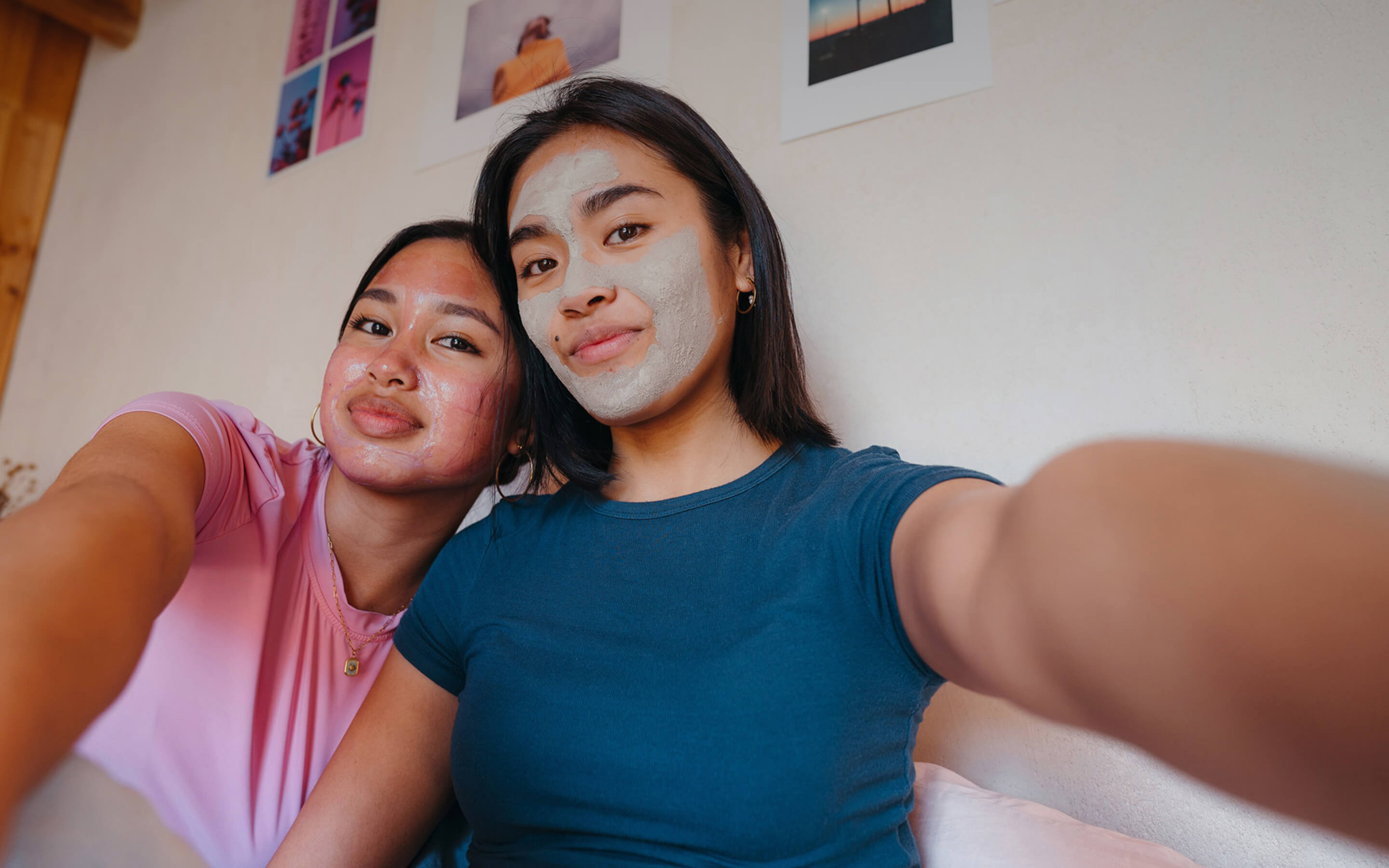 Two young women take a close-up selfie together indoors, both smiling softly while wearing facial masks—one with a pinkish gel mask and the other with a gray clay mask. They sit side by side against a cream-colored wall decorated with colorful art prints, creating a cozy, relaxed atmosphere that suggests a self-care or spa day at home.
