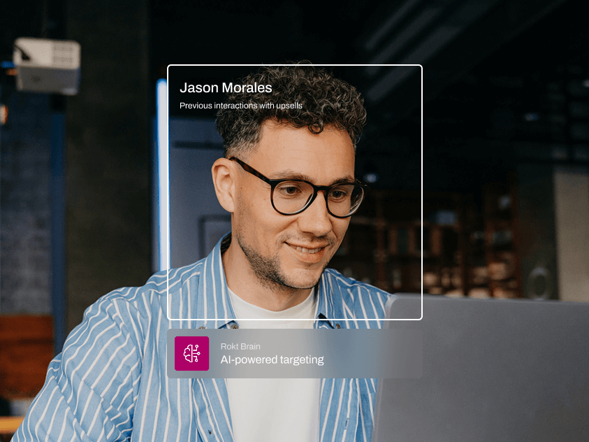 A man with curly hair and glasses smiles while looking at a screen, seated in a modern office space. A digital overlay highlights his name, “Jason Morales,” with a note that he has “Previous interactions with upsells.” Below that, a banner reads “Rokt Brain – AI-powered targeting,” suggesting a data-driven personalization experience.