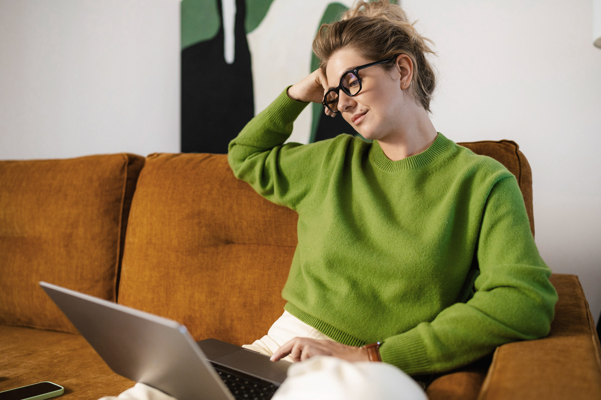 A woman in a bright green sweater and glasses sits on a brown couch, leaning her head on one hand while looking at a laptop. Her relaxed expression suggests she's thoughtfully engaged with something on the screen.