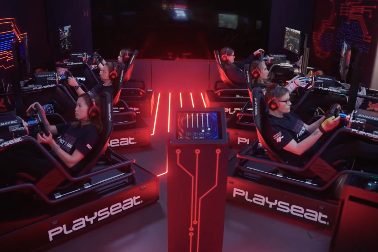 A row of competitive gamers, each seated in immersive racing simulator rigs branded “Playseat,” grips steering wheels and wears headsets. Red neon lighting and a central touchscreen control panel enhance the high-tech setup.