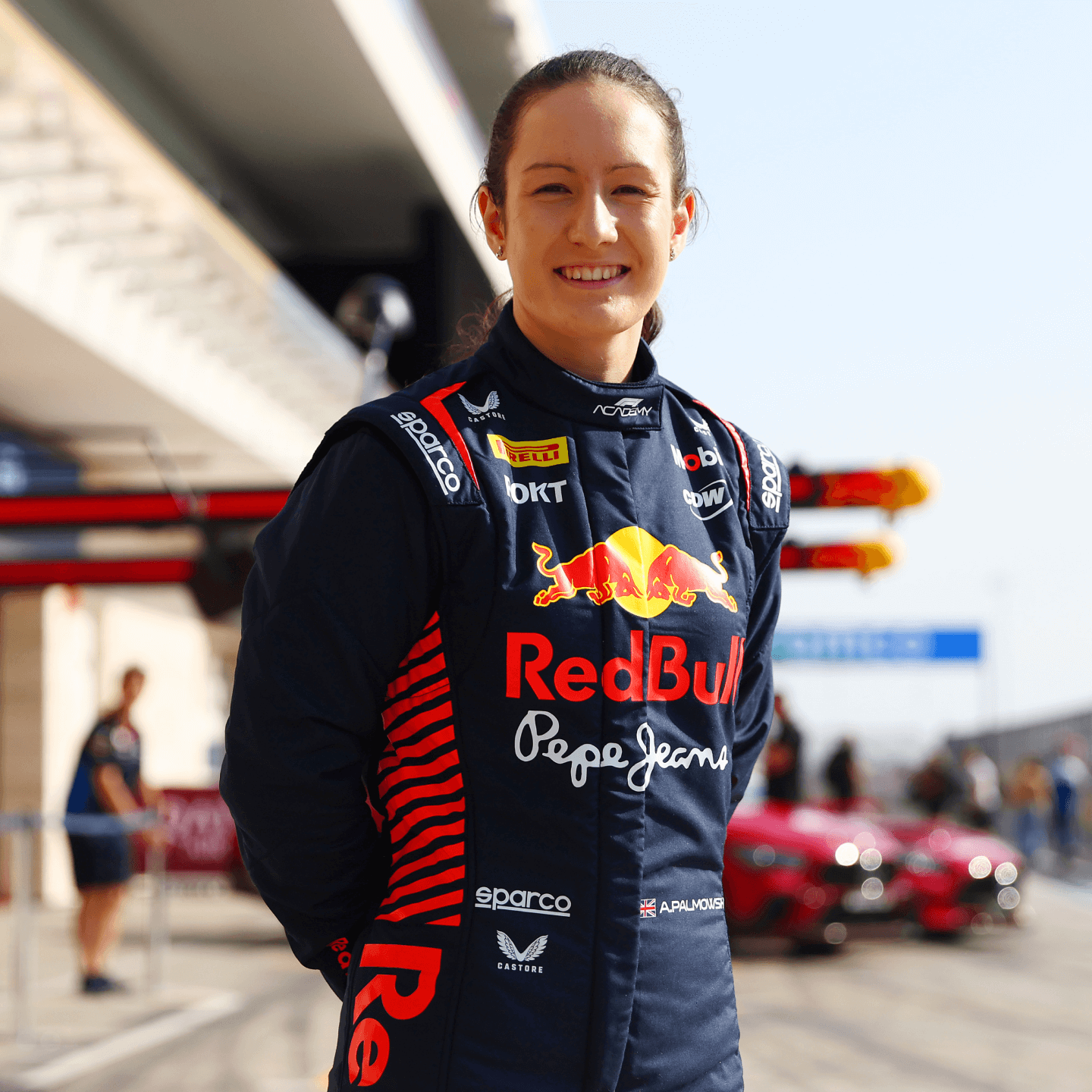 A female race car driver stands smiling in a Red Bull Racing fireproof suit at a Formula 1 paddock. The navy-blue suit features bold red and yellow sponsor logos including Red Bull, Pepe Jeans, and Pirelli, with red-striped detailing along the sides. Her dark hair is tied back neatly. Behind her, pit lane activity continues, with two red sports cars parked and crew members blurred in the background under bright daylight.