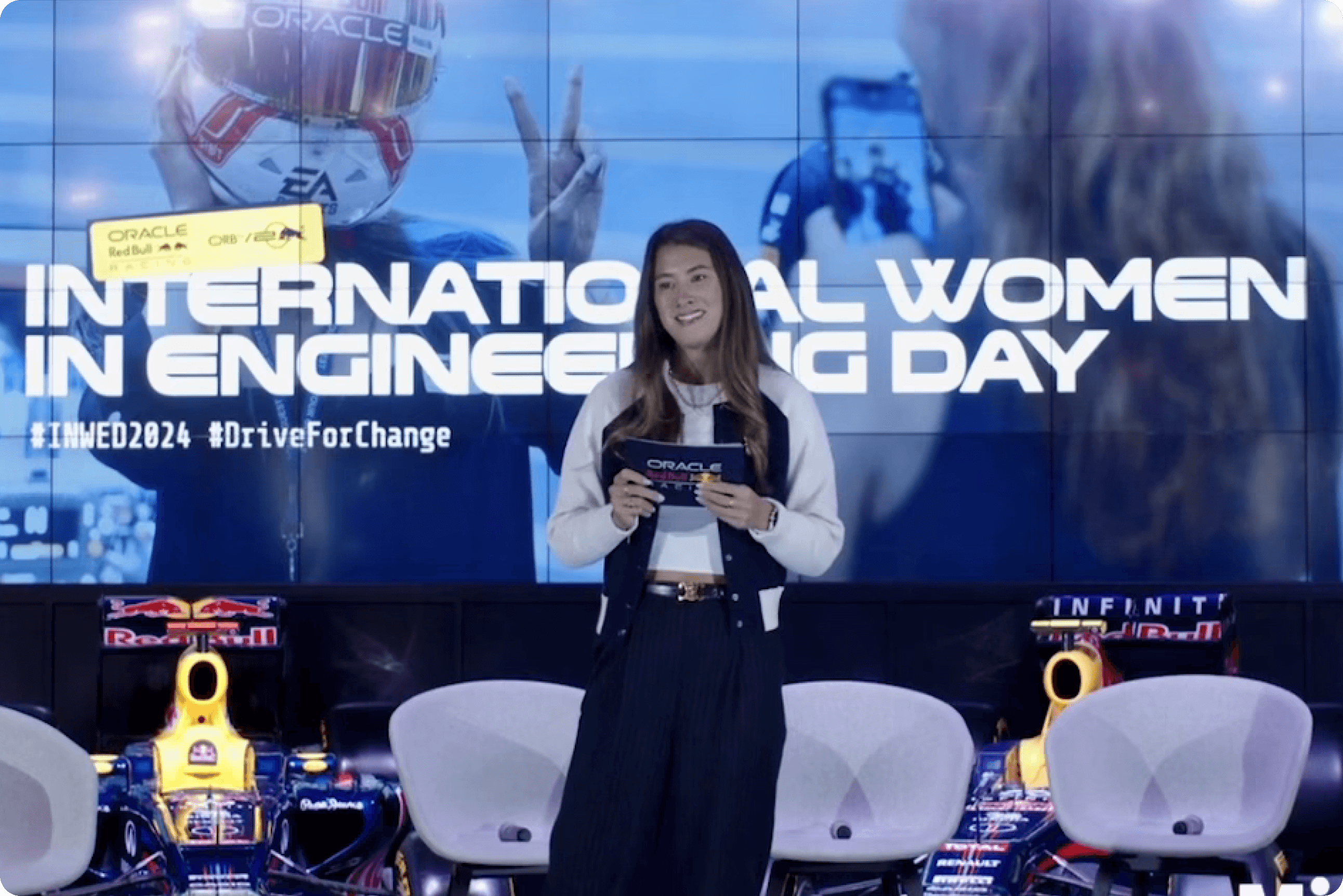 A woman stands on stage holding notes at an International Women in Engineering Day event, with Red Bull Racing F1 car nose sections and a large branded screen behind her.