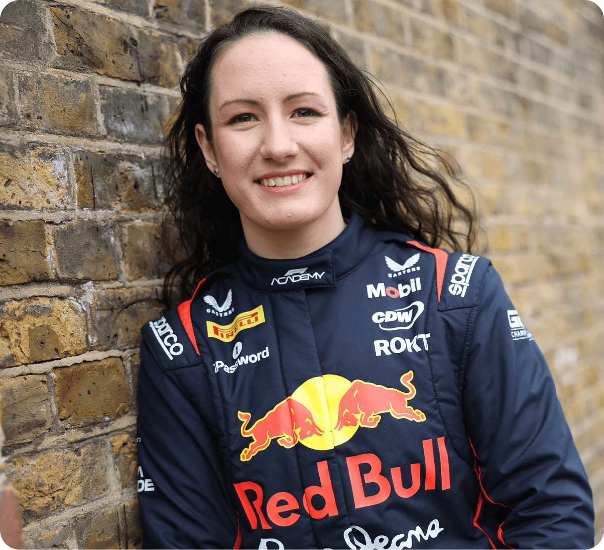 A woman in a Red Bull Racing fireproof suit smiles while leaning against a brick wall. She has long wavy dark hair and stands confidently, embodying a professional motorsport setting.