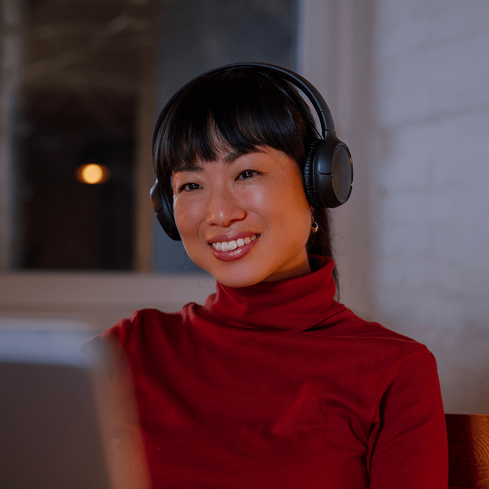 A woman wearing black over-ear headphones and a red turtleneck smiles warmly while looking at a laptop screen, likely engaged in a video call or virtual meeting. She is seated indoors in a softly lit room with a blurred window and brick wall in the background, creating a cozy, focused atmosphere. Her expression suggests attentiveness and enjoyment.