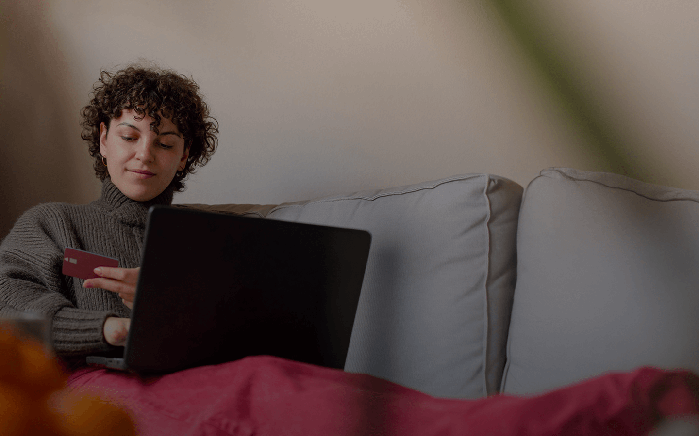 A person with short curly hair relaxes on a gray couch, holding a credit card in one hand while using a laptop on their lap. They wear a cozy gray turtleneck sweater and appear focused, likely making an online purchase. A red blanket covers their legs, adding to the casual and comfortable home setting.