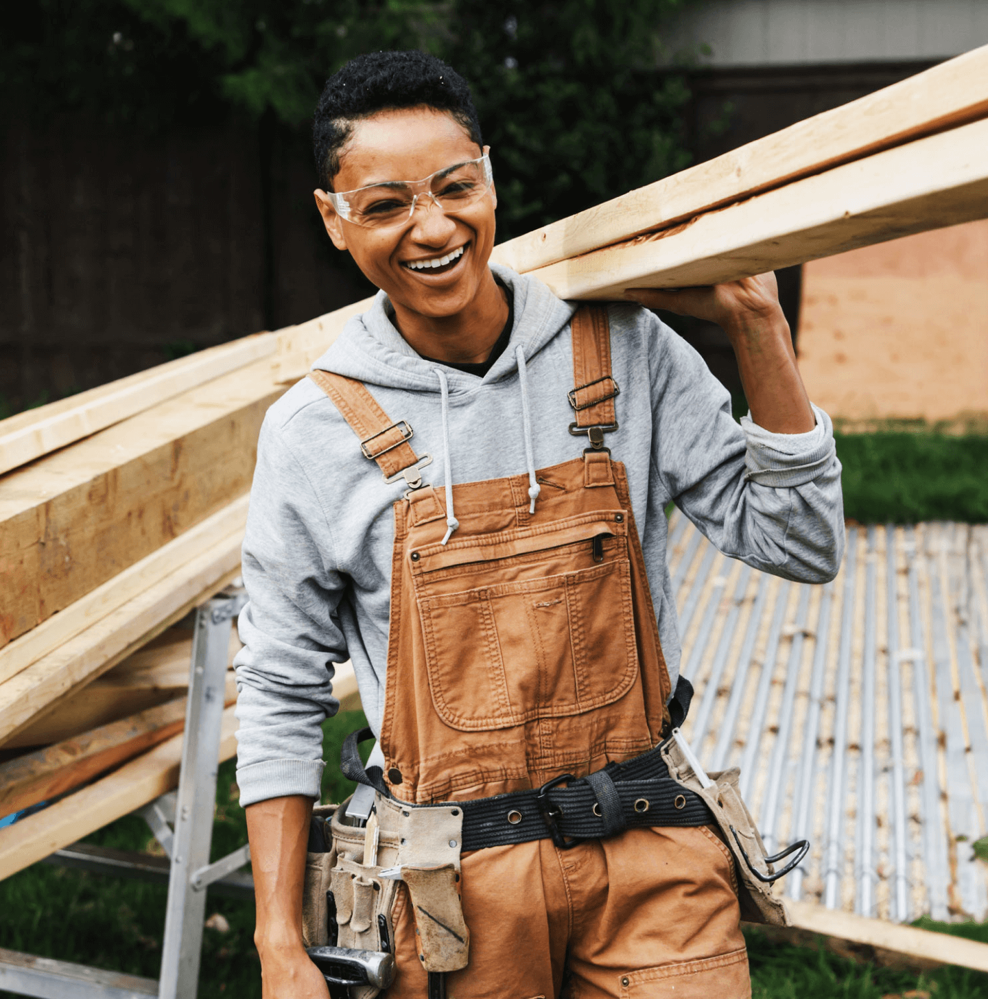 A smiling construction worker carries several long wooden beams on their shoulder at a job site. They wear a gray hoodie under tan overalls with a black tool belt and safety goggles. The background shows more lumber, a ladder, and a partially completed structure, suggesting active outdoor construction work. The worker appears confident and relaxed, adding a positive, human touch to the scene.