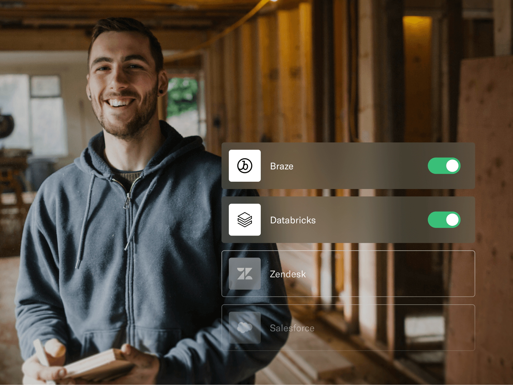 A smiling man in a dark hoodie stands in a partially renovated room, holding a notebook or tablet. He appears relaxed and confident, likely involved in construction or project planning. On the right side of the image, a digital interface overlay shows toggles for integrating software tools—Braze and Databricks are switched on, while Zendesk and Salesforce are grayed out. The image highlights a blend of hands-on craftsmanship with modern data and workflow management tools.