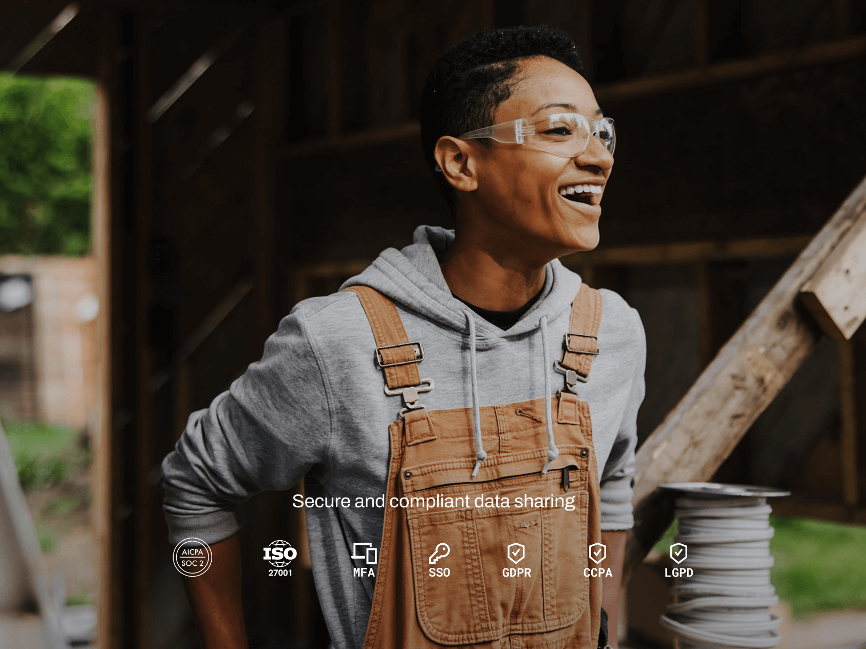 A person wearing safety glasses, a gray hoodie, and brown overalls stands smiling at a construction site, exuding confidence and enthusiasm. They are near a stack of coiled electrical wire, suggesting involvement in building or renovation work. Overlaid at the bottom are icons and text promoting “Secure and compliant data sharing,” with references to standards and regulations like SOC 2, ISO 27001, MFA, SSO, GDPR, CCPA, and LGPD—blending themes of cybersecurity with hands-on labor.