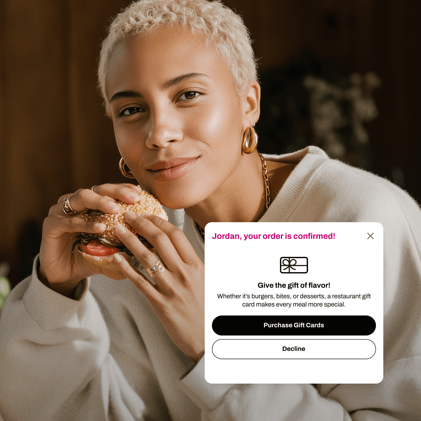 A woman with short platinum blonde hair and gold hoop earrings smiles softly while holding a fresh sandwich with lettuce, tomato, and sesame seed bun. She wears a cream-colored sweater and gold jewelry, sitting in a warmly lit space with blurred floral decor in the background. Overlaid on the image is a pop-up confirmation message that reads: “Jordan, your order is confirmed!” Below it, a call-to-action promotes restaurant gift cards with the text, “Give the gift of flavor! Whether it’s burgers, bites, or desserts, a restaurant gift card makes every meal more special.” Two buttons below offer options to “Purchase Gift Cards” or “Decline.”