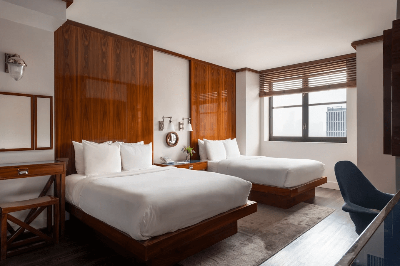 Modern hotel room with two wooden platform beds, white bedding, wooden wall panels, a window with blinds, and a dark chair.