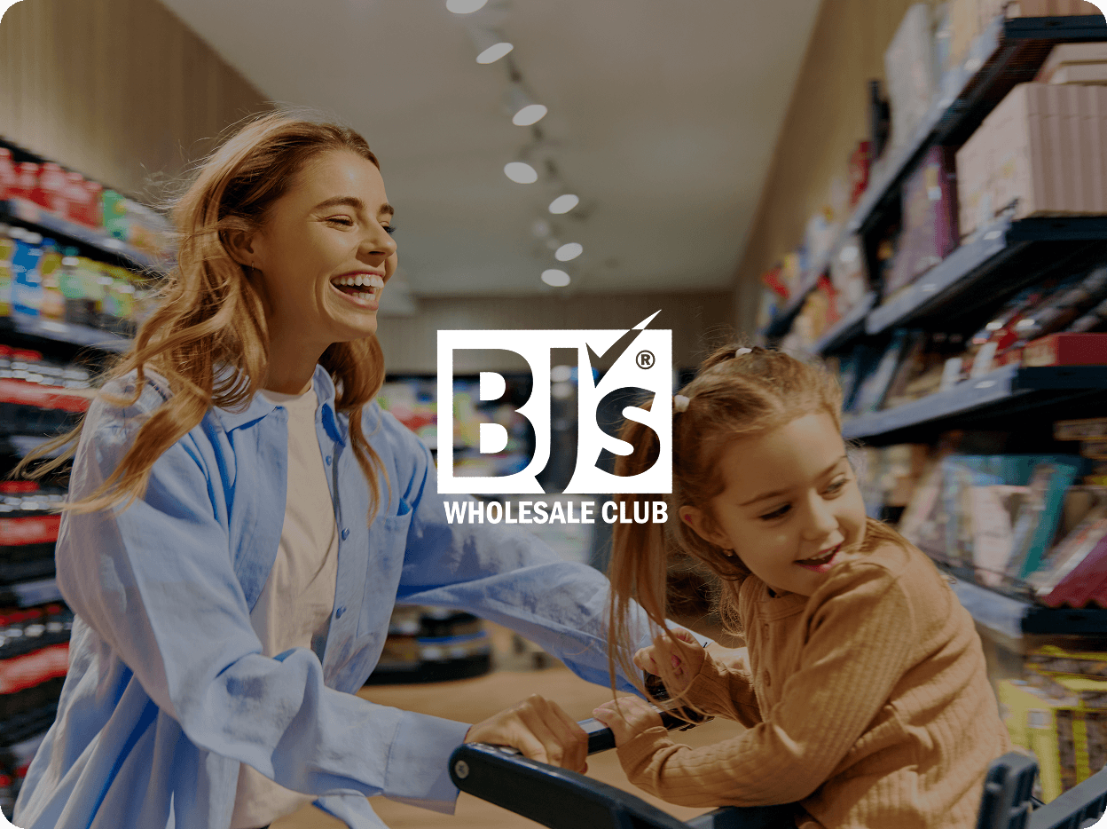 Smiling woman pushing a shopping cart with a young girl inside it in an aisle of a store with products on shelves.
