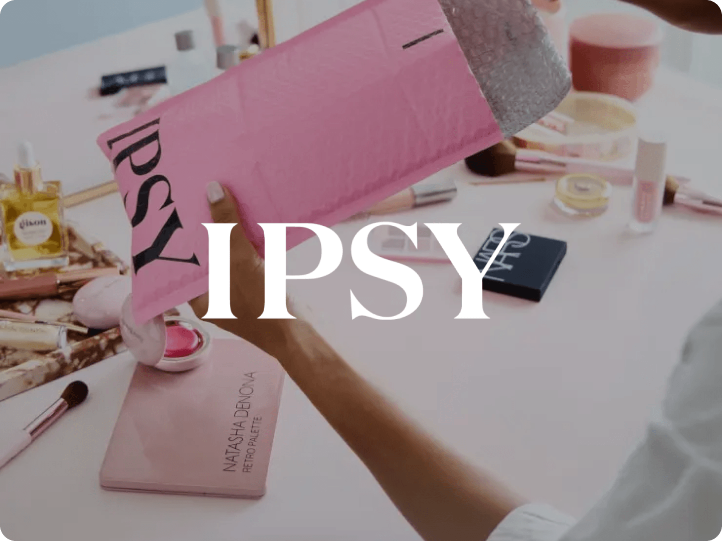 A person holding a pink IPSY padded mailer above a table with various makeup products including brushes, Natasha Denona Retro Palette, NARS compact, lip balm, and perfume bottle.