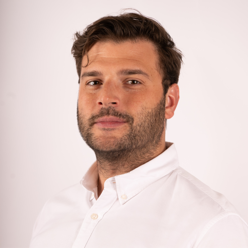 Headshot of a man with short dark hair and a beard wearing a white button-up shirt against a plain light background.
