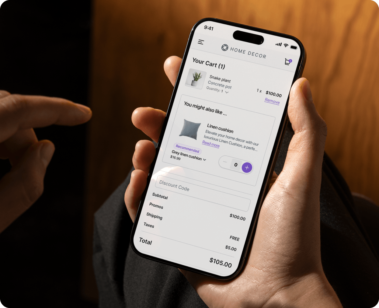 Hand holding a smartphone displaying an e-commerce shopping cart for 'Home Decor' with a snake plant priced at $100, a recommended grey linen cushion option for $16.99, and a total price of $105 including taxes and free shipping.