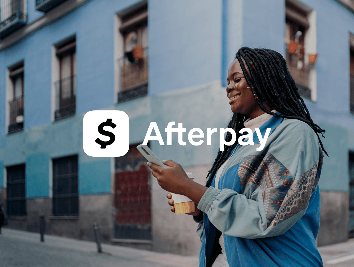 Smiling woman with long braided hair holding a smartphone and a takeout coffee cup walks on a street with blue buildings, with the Afterpay logo overlayed on the image.