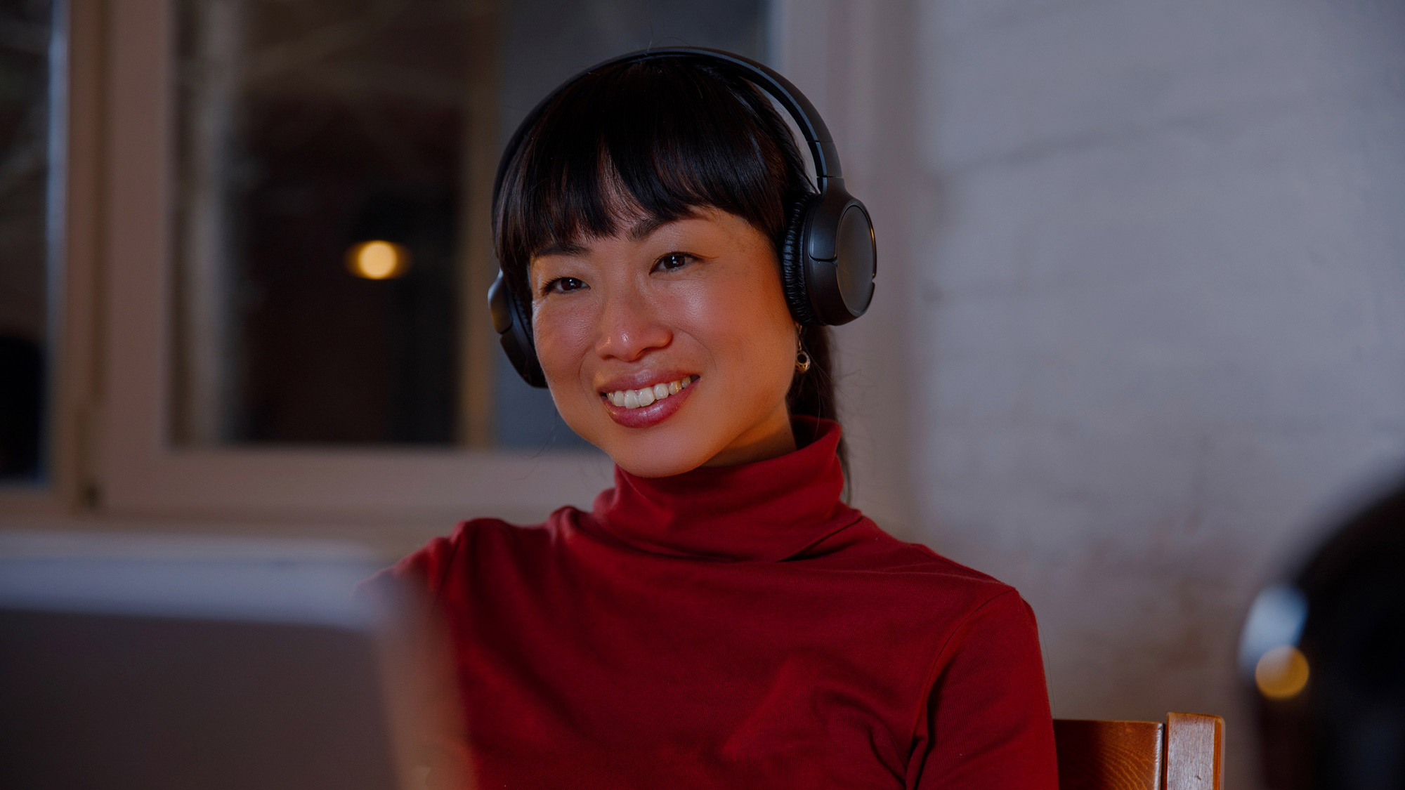 A woman wearing black over-ear headphones and a red turtleneck smiles warmly while looking at a screen, likely during a video call. She is seated indoors in a softly lit room with a blurred window in the background, creating a cozy and focused atmosphere.