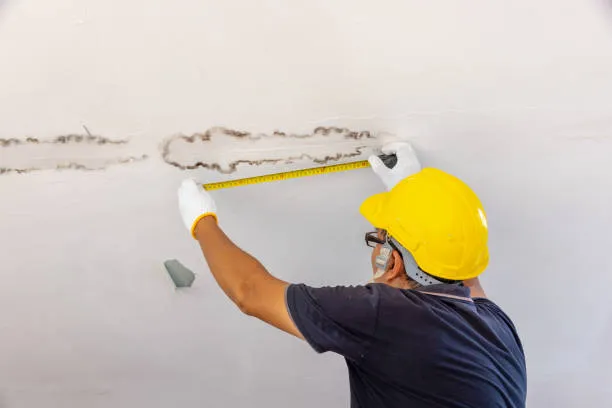 Worker in yellow hard hat measuring wall damage with measuring tape