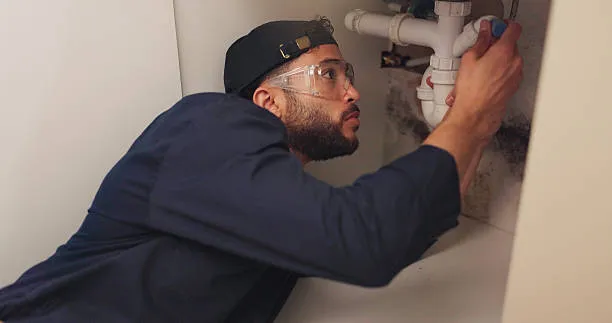 Plumber wearing safety glasses inspects pipes under sink