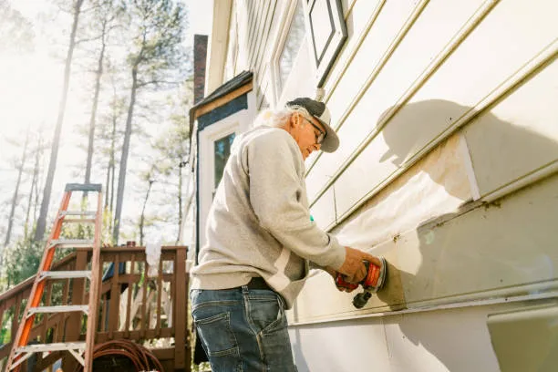Older worker using power drill on house exterior near ladder