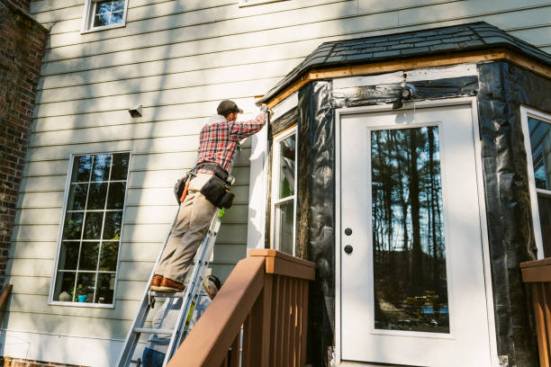 Worker painting house exterior on ladder near bay window and wooden deck