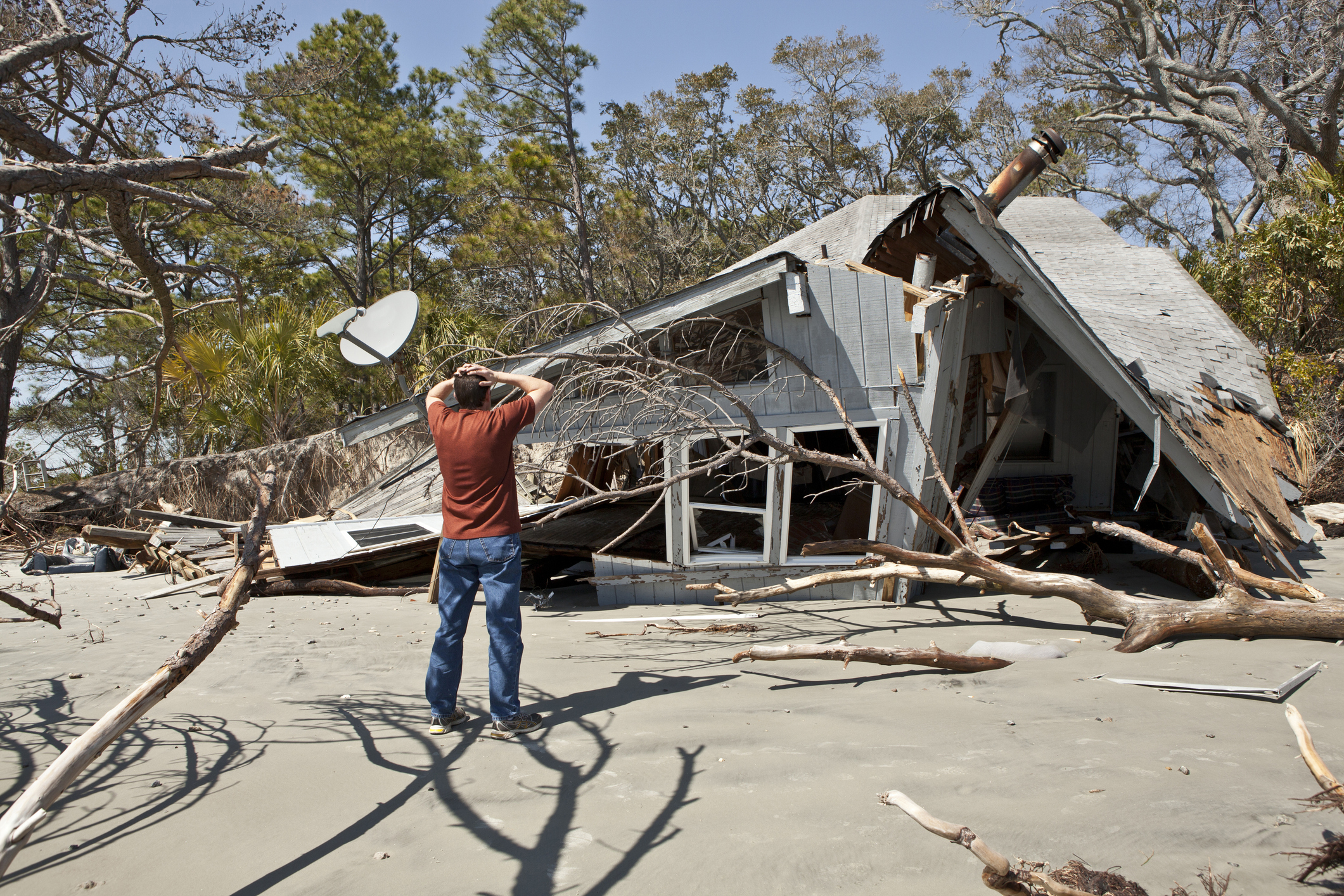Person surveys damaged house after hurricane, surrounded by debris and fallen trees