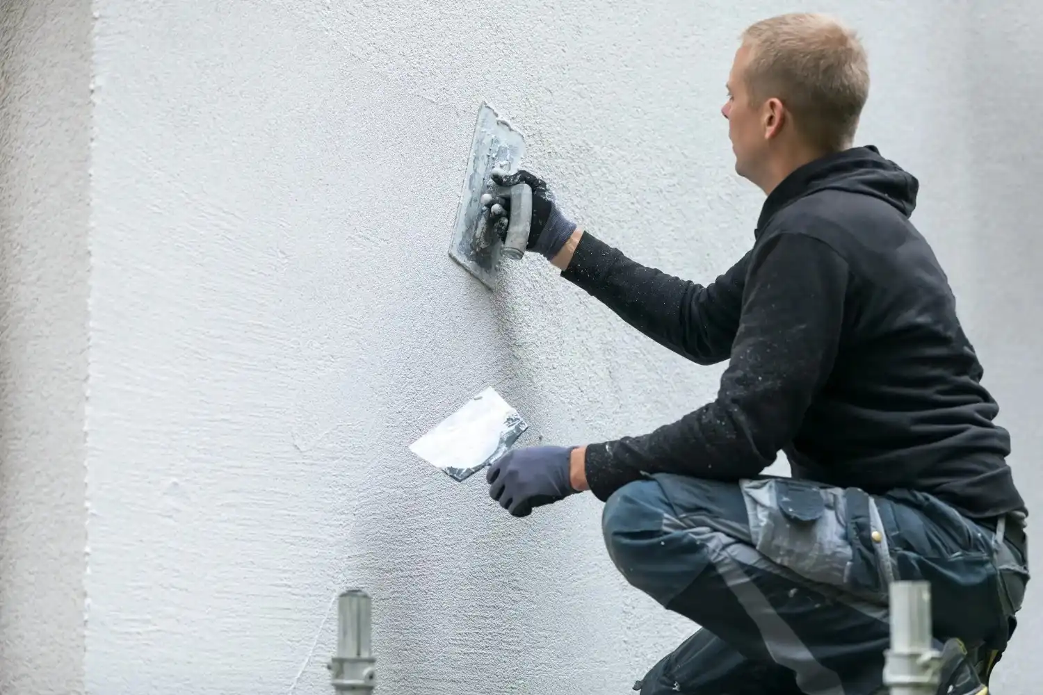 A man kneeling and applying a textured stucco finish to an exterior wall with a trowel.