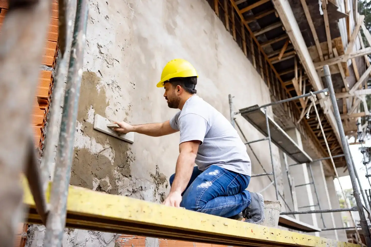 A construction worker on a scaffold, applying stucco to a brick wall with a trowel.