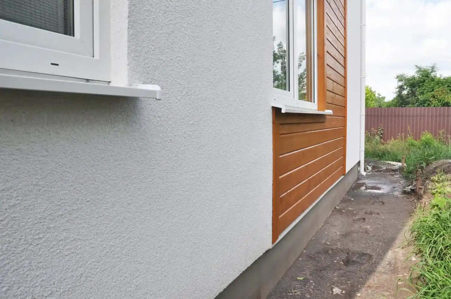 The exterior wall of a house, half covered in white textured stucco and half in brown wood siding.