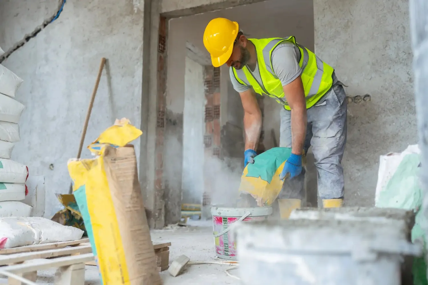 A construction worker in a hard hat and safety vest pouring dry cement mix from a bag into a bucket.