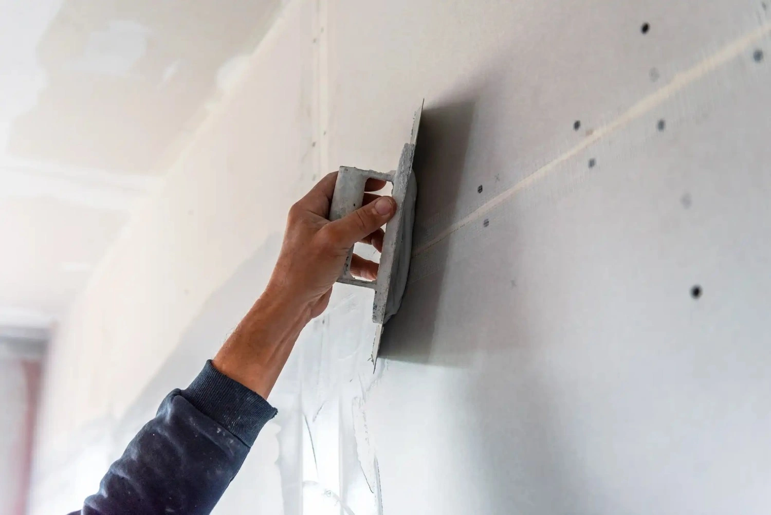 A hand holding a flat trowel and smoothing plaster over a wall with screws visible in the drywall.
