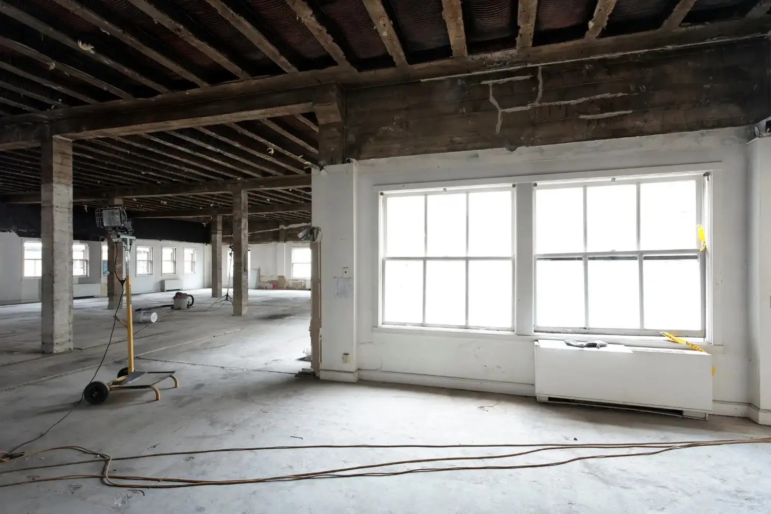 An empty, gutted room is shown from a low angle. The exposed wooden ceiling beams and a large window are visible. A yellow and black tripod with a light sits on the left side of the room.