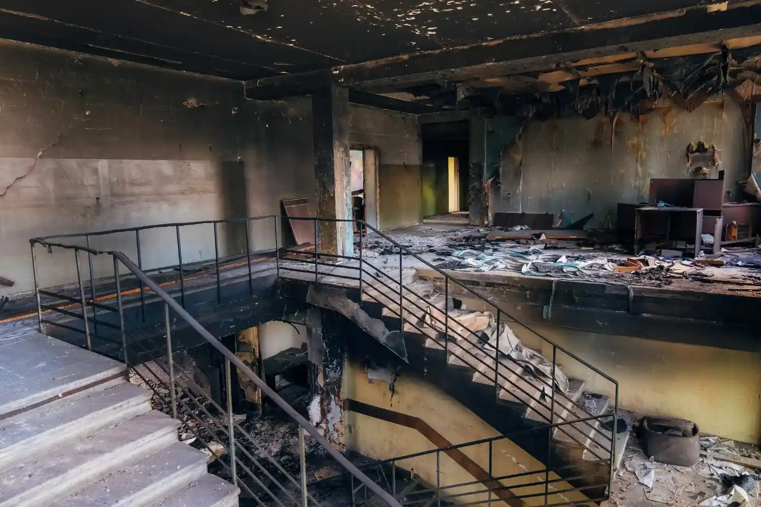 The interior of a fire-damaged building shows a scorched staircase with black railings, a destroyed ceiling, and debris scattered across the floor.